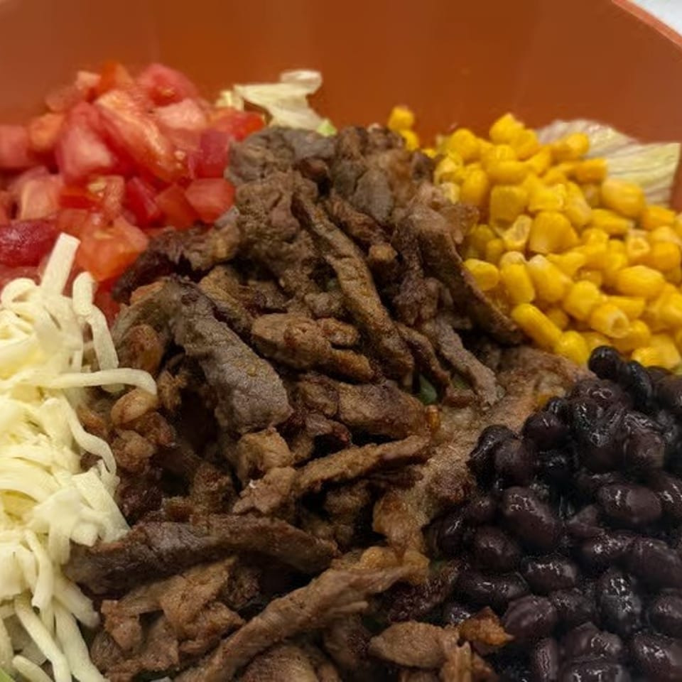 Bowl of seasoned grilled beef strips with black beans, corn, diced tomatoes, shredded cheese, and lettuce.