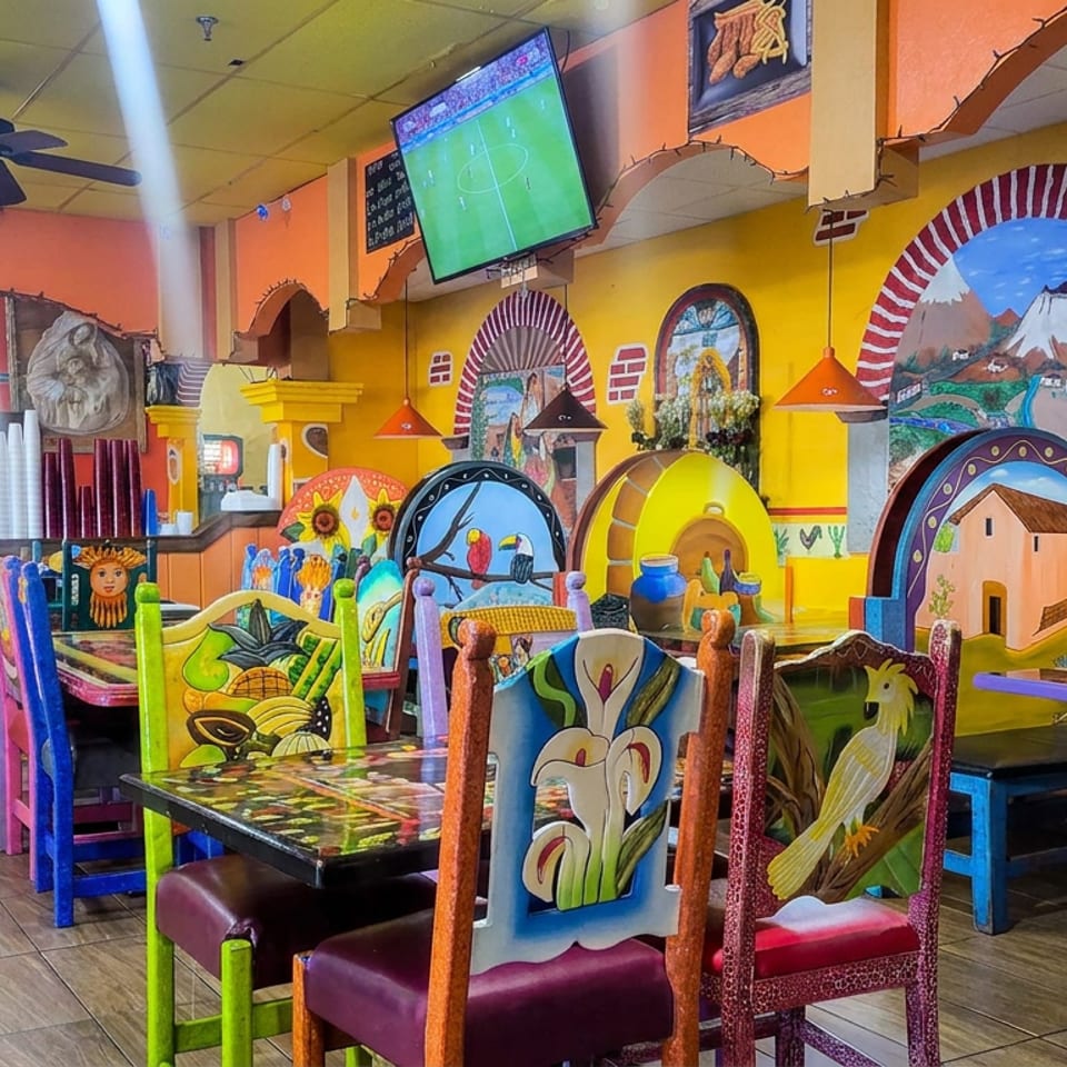 Colorful Mexican restaurant dining room with hand-painted chairs, murals, booths, pendant lights, and a television showing soccer.